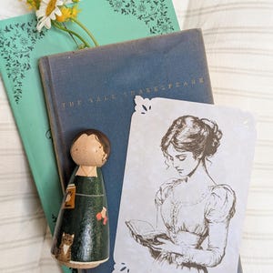 May include: A wooden peg doll in a green dress with a cat, a vintage book, a teal book with floral corner designs, a print of a woman reading, and a sprig of flowers. The book is titled "The Yale Shakespeare."