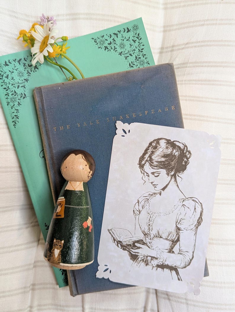 May include: A wooden peg doll in a green dress with a cat, a vintage book, a teal book with floral corner designs, a print of a woman reading, and a sprig of flowers. The book is titled "The Yale Shakespeare."