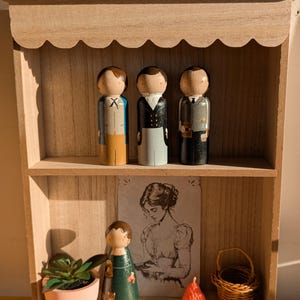 May include: Wooden shelf with three painted peg dolls dressed in suits. A small succulent in a pink pot, a drawing of a woman, a peg doll in a green dress, a small wicker basket, and a red strawberry-shaped object are also on the shelf.