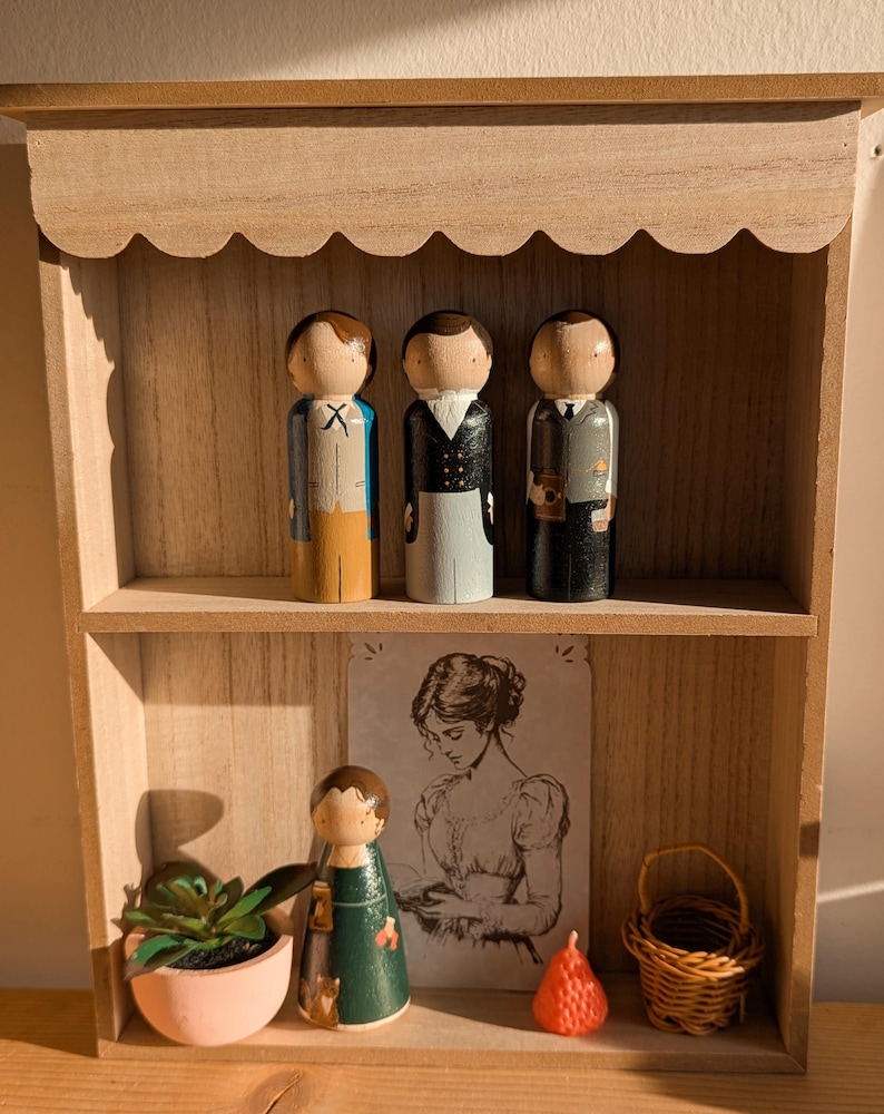 May include: Wooden shelf with three painted peg dolls dressed in suits. A small succulent in a pink pot, a drawing of a woman, a peg doll in a green dress, a small wicker basket, and a red strawberry-shaped object are also on the shelf.