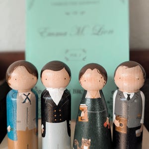 May include: Four wooden peg dolls, each painted to resemble characters, stand in front of a book. The dolls are dressed in various outfits, including a blue coat, black jacket, and green dress. The book has the title "Emma M. Lion" on the cover.