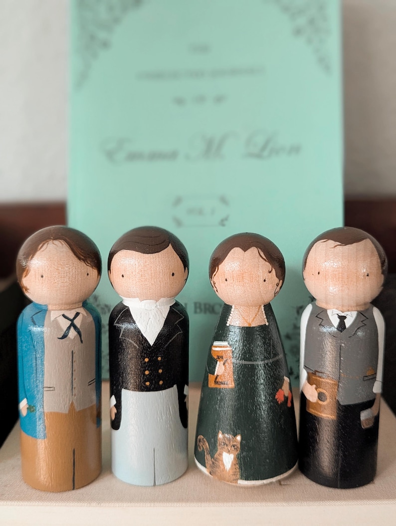 May include: Four wooden peg dolls, each painted to resemble characters, stand in front of a book. The dolls are dressed in various outfits, including a blue coat, black jacket, and green dress. The book has the title "Emma M. Lion" on the cover.
