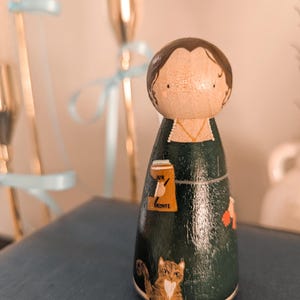 May include: A hand-painted wooden peg doll with a dark green dress and brown hair. The doll features a cat, a book, and a beer illustration. The text "Jane Bronte" is on the doll's dress.