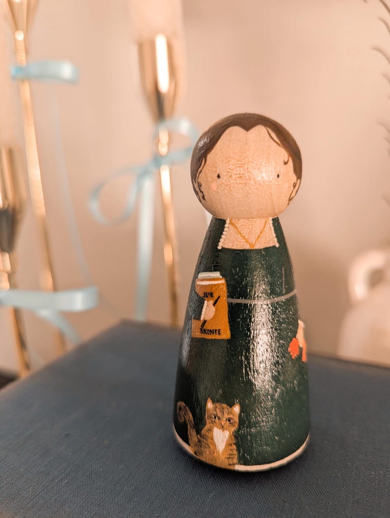 May include: A hand-painted wooden peg doll with a dark green dress and brown hair. The doll features a cat, a book, and a beer illustration. The text "Jane Bronte" is on the doll's dress.