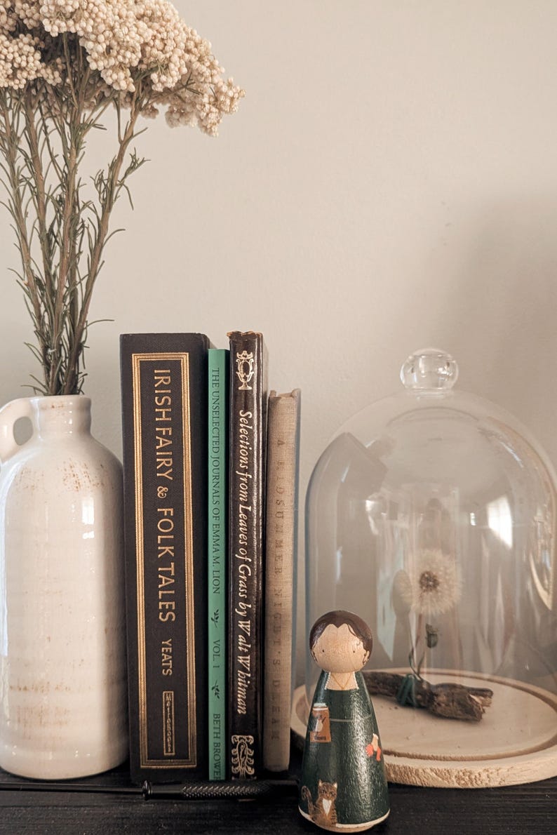 May include: A still life arrangement featuring a white ceramic jug with dried flowers, vintage books, and a glass cloche. A wooden peg doll stands in front of the cloche. The books include "Irish Fairy & Folk Tales" and "Selections from Leaves of Grass."