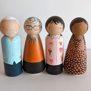 May include: Four wooden peg dolls, each painted with unique details. The first doll has white hair and a blue shirt, the second has brown hair and a colorful orange shirt, the third has black hair and a pink shirt with ice cream designs, and the fourth has brown hair and a black and orange floral dress.