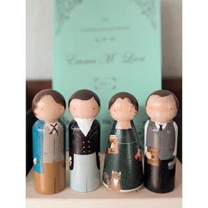 May include: Four wooden peg dolls, each with a painted face and clothing. The dolls are in the style of characters from a book, with one doll wearing a blue coat and another a black coat. A book with the title "Emma M. Lion" is in the background.