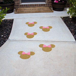 May include: A concrete walkway with five gold glitter Minnie Mouse head decals with pink bows. The decals are spaced out along the walkway, leading to a door with a welcome mat.