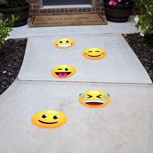 May include: Five yellow emoji stickers on a gray concrete walkway. The emojis are smiling, winking, sticking out its tongue, crying, and laughing.