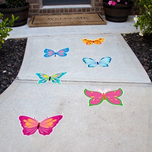May include: A concrete walkway with six colorful butterfly decals. The butterflies are pink, blue, yellow, green, and orange. The walkway is surrounded by green grass and bushes.