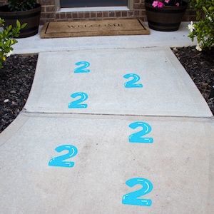May include: A concrete walkway with six blue numbers "2" painted on it. The numbers are in a line, leading to a door.