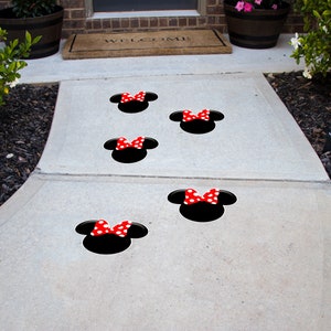 May include: A concrete walkway with five black and red Minnie Mouse head decals. The decals are spaced out in a line, leading up to a door.