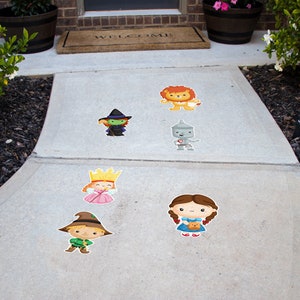 May include: A collection of colorful, cartoon-style character stickers from The Wizard of Oz, including Dorothy, the Lion, the Tin Man, the Wicked Witch, the Scarecrow, and a princess. The stickers are arranged on a concrete walkway.