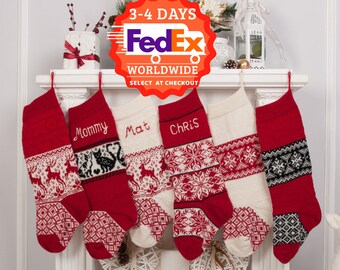 May include: Four red and white knitted Christmas stockings with personalized names "Mommy", "Mat", "Chris" and a fourth stocking with a snowflake pattern hanging on a white mantel. The stockings are decorated with a reindeer and snowflake pattern. A white deer figurine is on the right side of the image. A FedEx logo with the text "3-4 Days Worldwide Select at Checkout" is in the top center of the image.