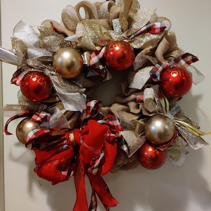 Könnte beinhalten: Ein festlicher Weihnachtskranz mit einer Basis aus Jute, verziert mit goldenen und silbernen Bändern, roten und goldenen Kugeln und einer großen roten Schottenkaro-Schleife. Die Kugeln tragen die Wörter "CHRIST" und "ATLAS".