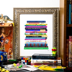 May include: A framed print of a stack of colorful paperback books, including titles like "The Giving", "Matilda", "The Phantom Tollbooth", and "The Writing Game". The print is on a white background and is surrounded by a gold frame. There are other items in the image, including a toy dinosaur, a doll, a mug, and a Rubik's Cube.