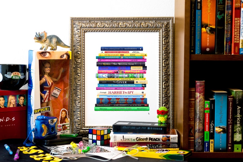 May include: A framed print of a stack of colorful paperback books, including titles like "The Giving", "Matilda", "The Phantom Tollbooth", and "The Writing Game". The print is on a white background and is surrounded by a gold frame. There are other items in the image, including a toy dinosaur, a doll, a mug, and a Rubik's Cube.