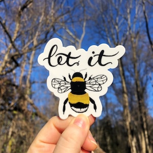 Puede incluir: Una pegatina blanca con una ilustración de abeja y las palabras "let it" en escritura negra. La abeja tiene rayas amarillas y negras y alas detalladas. La pegatina se sostiene contra un fondo de árboles desnudos y un cielo azul.
