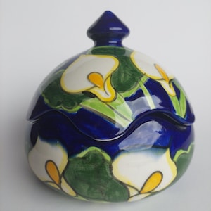 Op de afbeelding: Een keramische pot met deksel met een afgeronde vorm. De pot is beschilderd met een donkerblauwe basis, groene bladeren en witte calla lelies met gele centra. Het deksel is donkerblauw en puntig.