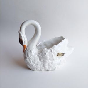Op de afbeelding: Een witte keramische zwaan planter met een gebogen nek en open bovenkant. De zwaan heeft getextureerde veren en een bruine snavel. Een klein gouden label is bevestigd. Ideaal voor kleine planten of bloemen.