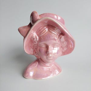 Pode incluir: Busto de cerâmica rosa de uma pessoa usando um chapéu de abas largas com um laço. O busto tem um acabamento brilhante e está sobre um fundo simples. A escultura é um item decorativo.
