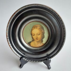 Può includere: Un piccolo ritratto rotondo incorniciato di una donna con un'aureola, posto su un cavalletto nero. Il ritratto presenta una donna con la pelle chiara, capelli biondi e un'espressione serena. La cornice è marrone scuro con un aspetto strutturato.