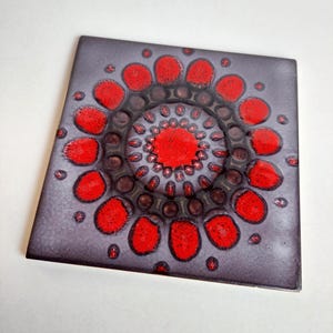 Könnte beinhalten: Quadratische Keramikfliese mit floralem Design. Die Fliese hat einen dunkelgrauen Hintergrund mit einem zentralen roten Kreis, der von roten, blütenblattförmigen Designs umgeben ist. Die Fliese hat eine glänzende Oberfläche.