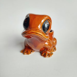 Può includere: Statuetta a forma di rana in ceramica marrone lucido, con grandi occhi neri. La rana è seduta, con le zampe palmate visibili. Il design della statuetta è stravagante e di tipo cartoon.