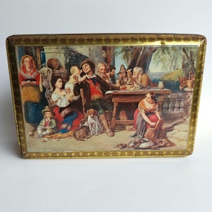 Può includere: Scatola rettangolare vintage con una pittura dettagliata di un raduno di persone, cani e un bambino. La scena è ambientata all'aperto con un tavolo e varie figure in abiti d'epoca. La scatola ha un bordo dorato.