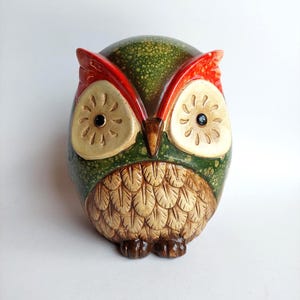 Może przedstawiać: Ceramiczna figurka sowy w kolorze zielonym i czerwonym. Sowa ma duże, okrągłe oczy, szczegółowy wzór piór na piersi i małe brązowe stopy. Dekoracyjny element.