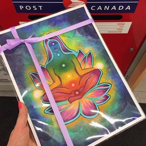 May include: A colorful print of a woman meditating in a lotus flower with a galaxy background. The woman is surrounded by a rainbow of colors representing the seven chakras. The print is tied with a purple ribbon.