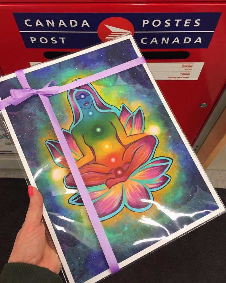 May include: A colorful print of a woman meditating in a lotus flower with a galaxy background. The woman is surrounded by a rainbow of colors representing the seven chakras. The print is tied with a purple ribbon.