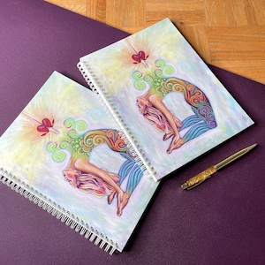 May include: Two spiral bound notebooks with colorful illustrations of a woman in a yoga pose. The notebooks have a white background with a light yellow and pink gradient. The woman is depicted with a colorful mandala design on her body. The notebooks are on a purple surface.