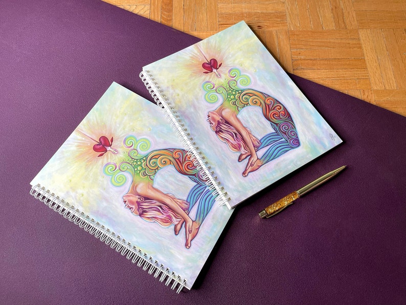 May include: Two spiral bound notebooks with colorful illustrations of a woman in a yoga pose. The notebooks have a white background with a light yellow and pink gradient. The woman is depicted with a colorful mandala design on her body. The notebooks are on a purple surface.