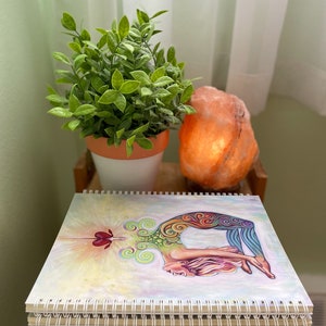 May include: A stack of spiral bound notebooks with a colorful illustration of a woman in a yoga pose on the front cover. The illustration features a rainbow color scheme and a heart shape in the center.