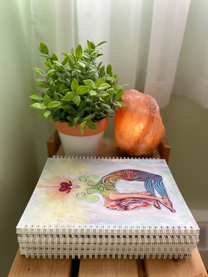 May include: A stack of spiral bound notebooks with a colorful illustration of a woman in a yoga pose on the front cover. The illustration features a rainbow color scheme and a heart shape in the center.