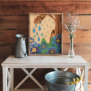 Puede incluir: Una pintura enmarcada de una mano que se extiende hacia abajo para tocar un campo de flores azules y moradas. La mano est&aacute; goteando gotas de lluvia azules. El fondo es una veta de madera clara.