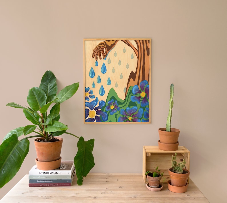 Puede incluir: Una pintura enmarcada que representa una mano vertiendo gotas de lluvia azules sobre un campo de flores azules y amarillas. El fondo es de madera marr&oacute;n claro.