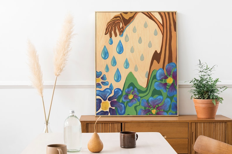 Puede incluir: Una pintura enmarcada que representa una mano que se extiende hacia abajo y libera gotas de lluvia azules sobre un campo de flores azules estilizadas con centros amarillos. El fondo es una veta de madera clara.