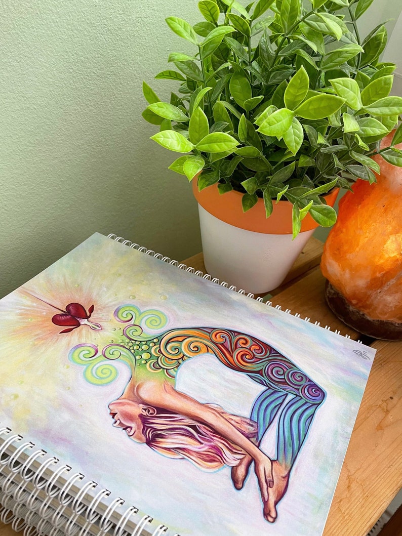 May include: A spiral bound sketchbook with a colorful illustration of a woman in a yoga pose. The illustration is done in a vibrant style with swirls and patterns. The background is a soft watercolor wash of light blue and pink.