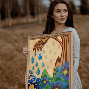 Puede incluir: Una mujer sostiene una pintura enmarcada con una mano que se extiende hacia abajo y gotas de lluvia azules que caen. La pintura tiene un dise&ntilde;o floral verde, azul y morado.