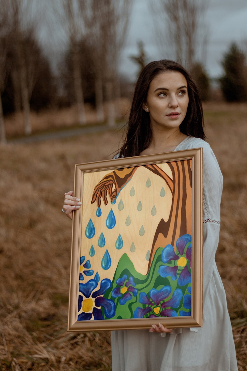 Puede incluir: Una mujer sostiene una pintura enmarcada con una mano que se extiende hacia abajo y gotas de lluvia azules que caen. La pintura tiene un dise&ntilde;o floral verde, azul y morado.