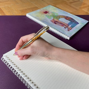 May include: A hand writing in a spiral bound notebook with a gold pen. The notebook is on a purple surface and a closed spiral bound notebook with a colorful illustration of a woman on the cover is behind it.