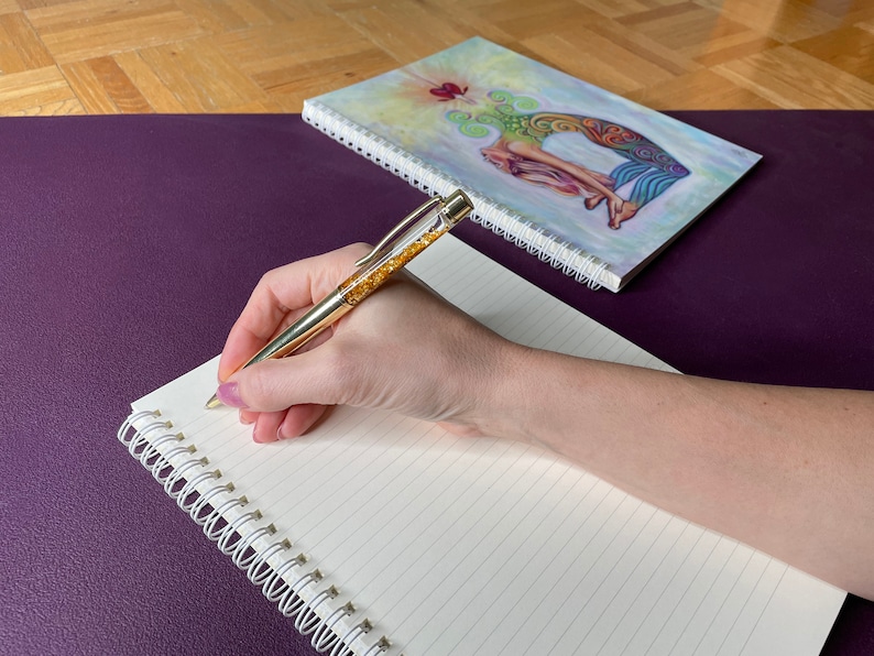 May include: A hand writing in a spiral bound notebook with a gold pen. The notebook is on a purple surface and a closed spiral bound notebook with a colorful illustration of a woman on the cover is behind it.