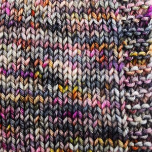 May include: Close-up of a knitted fabric with a variegated yarn in shades of grey, pink, orange, and yellow. The fabric is knit in a simple stitch pattern.