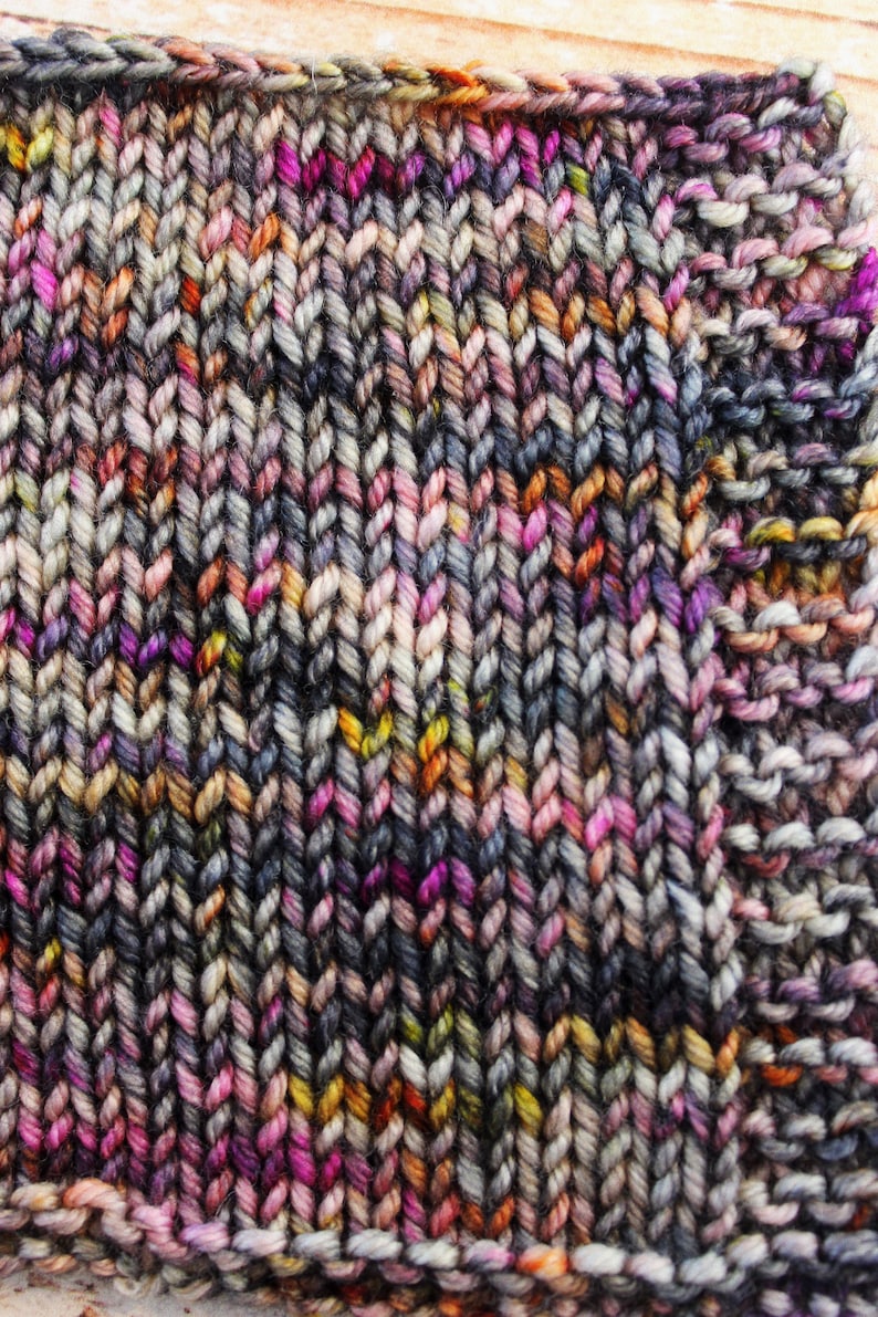 May include: Close-up of a knitted fabric with a variegated yarn in shades of grey, pink, orange, and yellow. The fabric is knit in a simple stitch pattern.