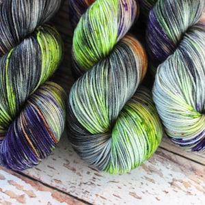 May include: Three skeins of yarn in a green, purple, and gray color scheme. The yarn is hand-dyed and has a variegated pattern.