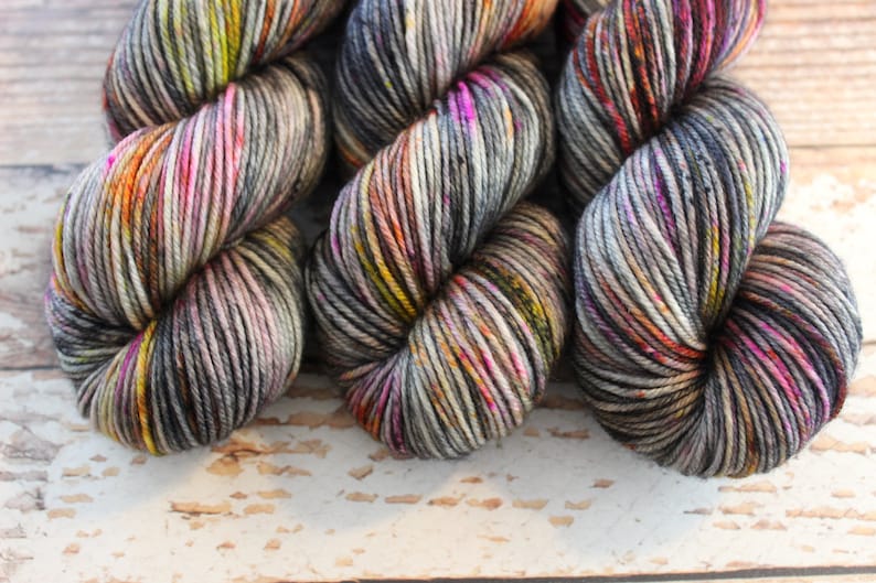 May include: Three skeins of yarn in a variegated colorway with shades of gray, black, orange, yellow, pink, and white.