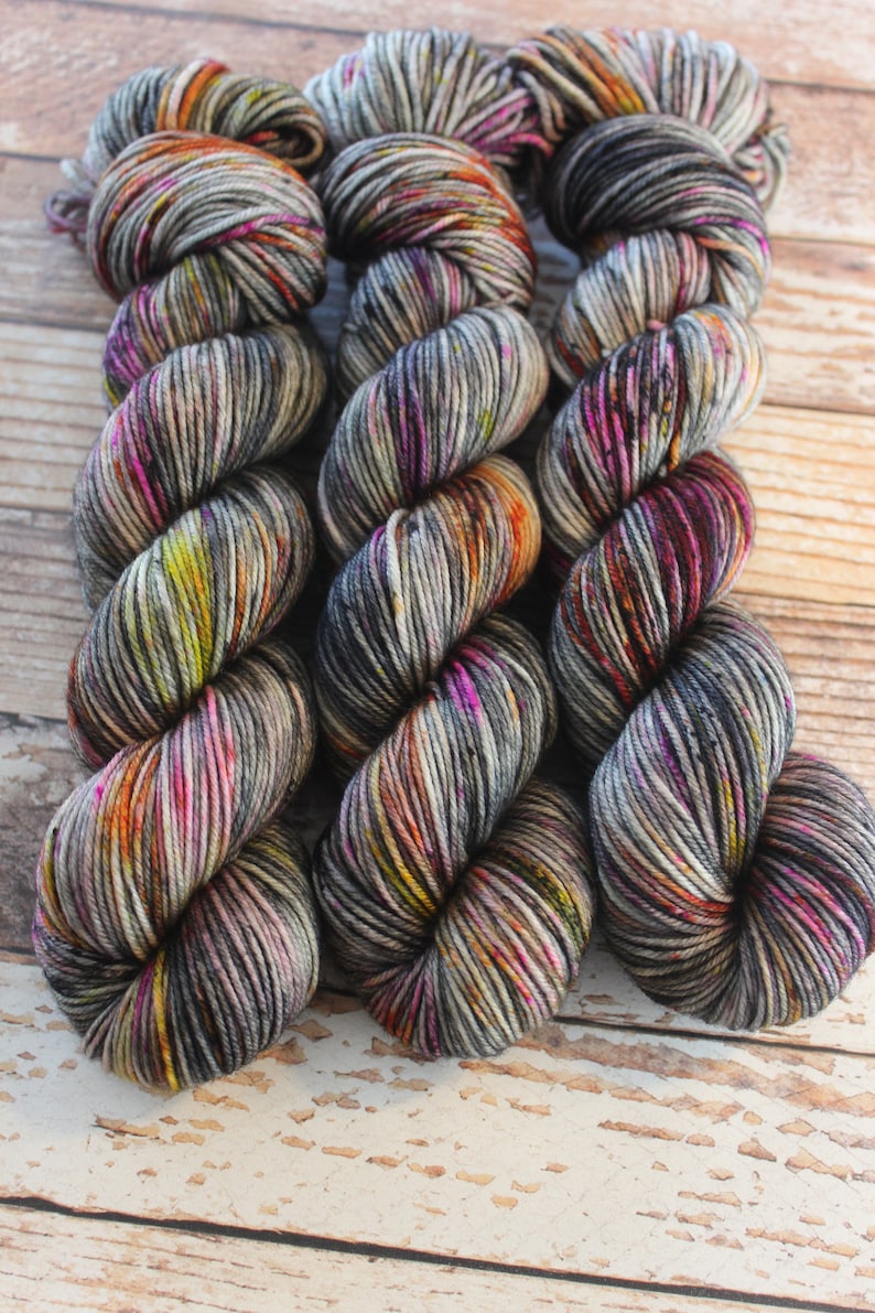 May include: Three skeins of colorful yarn in shades of grey, pink, orange, and yellow. The yarn is arranged in a circle, with the ends of the skeins overlapping.