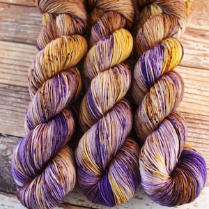 May include: Three skeins of yarn in a purple and brown colourway. The yarn is a variegated blend of purple, brown, and cream.